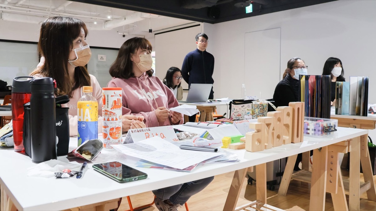 Teacher-training session: trainee teachers around long tables set with wooden number blocks, picture books and name cards, with an instructor leading from the front.