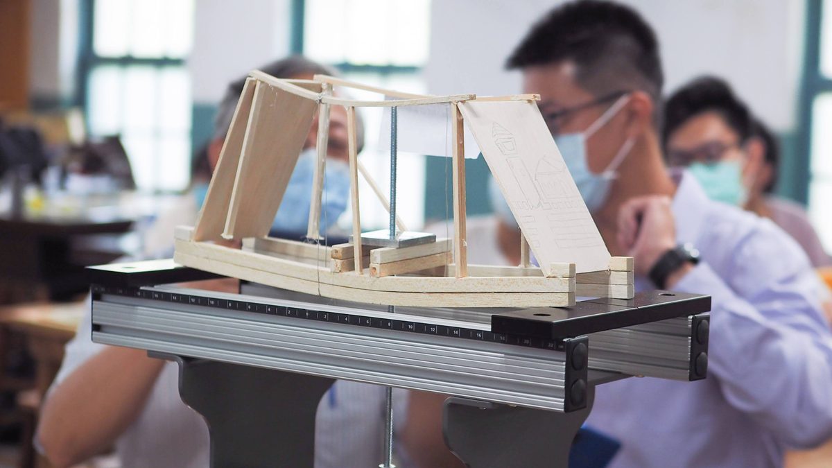Programme session: a participant-built wooden bridge model mounted on a metal test rig, with students and instructors gathered behind it in discussion.