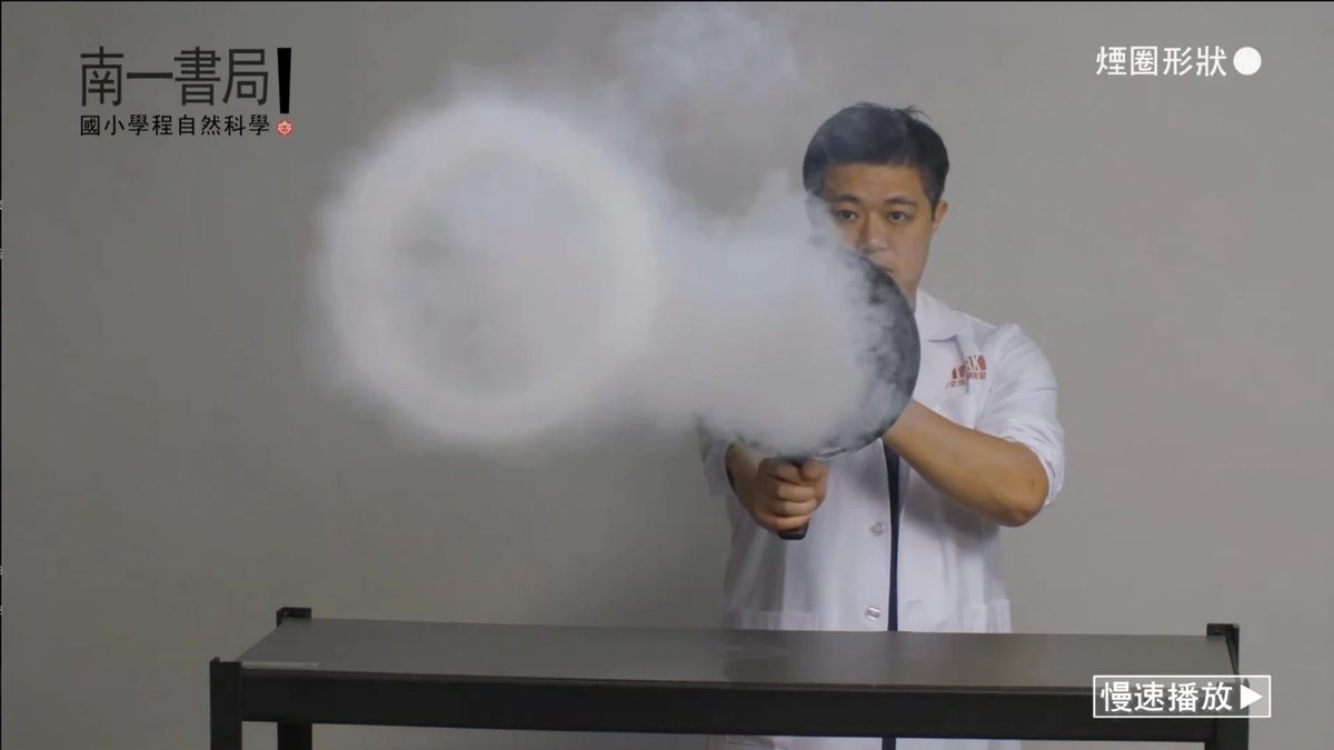 Frame from a textbook demonstration video: an instructor in a lab coat fires an air cannon, sending a clean smoke ring across the frame, with the publisher’s series title at top left.