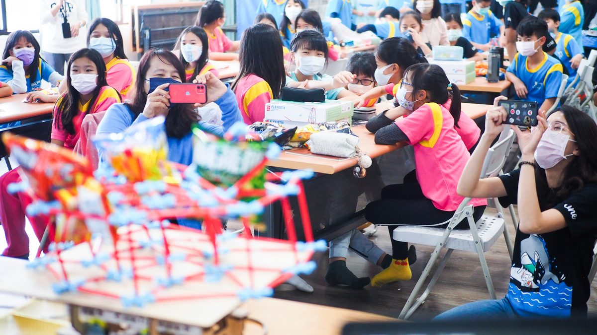 Primary-school session: a room full of pupils holding up phones to photograph a red and blue structural model on the table, with a wooden structural kit in the foreground.