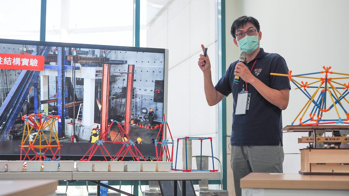 Science festival session: a speaker with a microphone explains a long table of red and blue structural models and wooden building kits, with footage of a full-scale structural test on the screen behind.