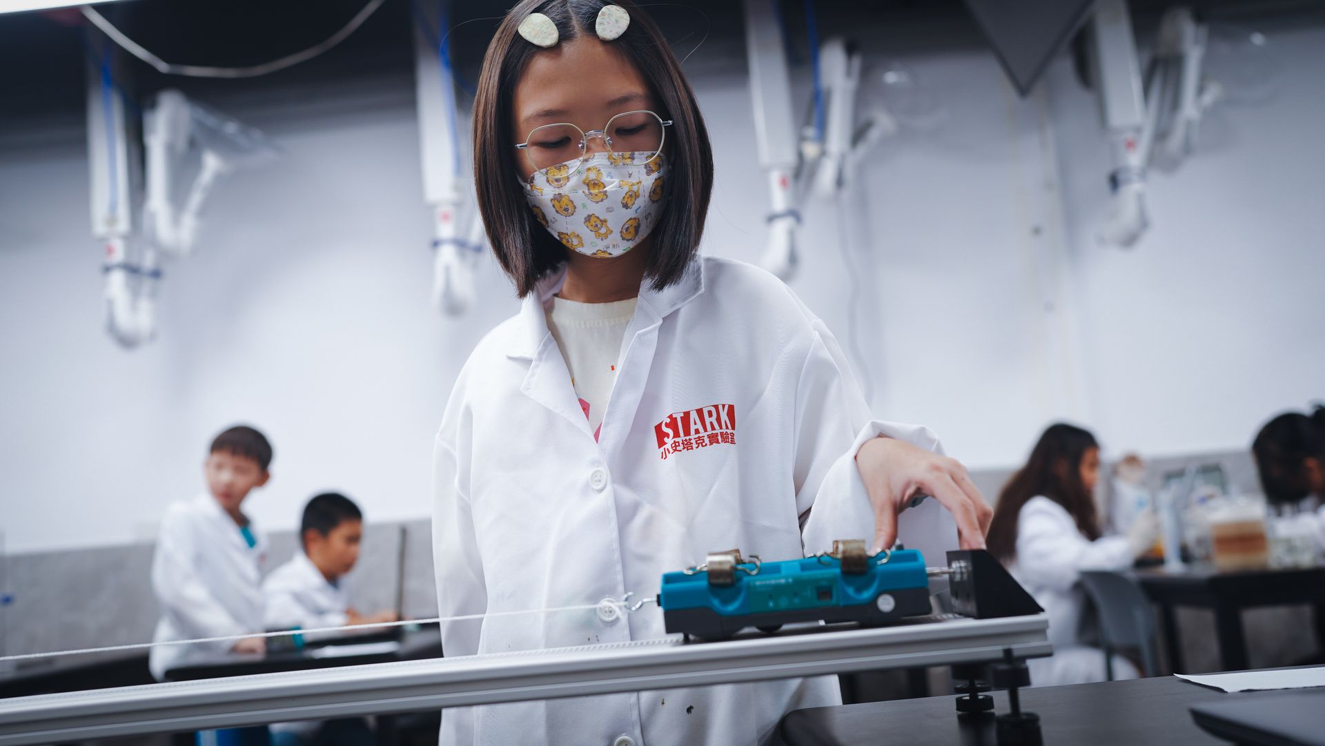 A girl in a STARK lab coat operating a dynamics cart on its track, other children at work behind her.