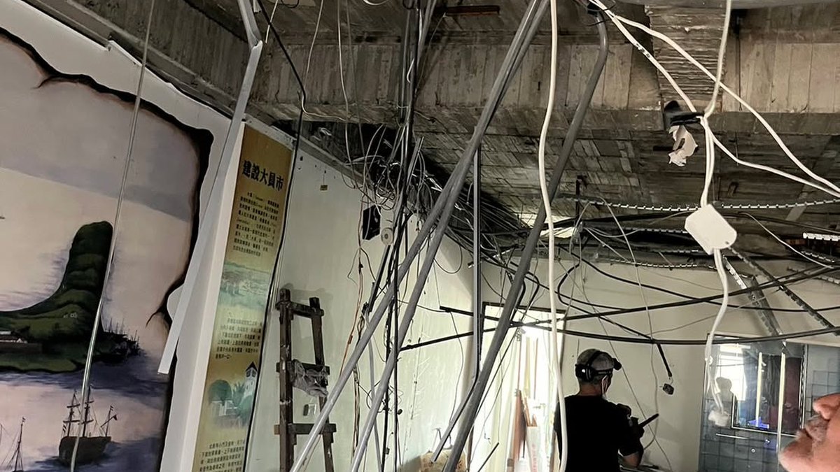 A classroom mid-renovation: the ceiling stripped back to bare concrete with bundles of cable hanging down, a masked worker at the far end, and a large mural of sailing ships on the left-hand wall.
