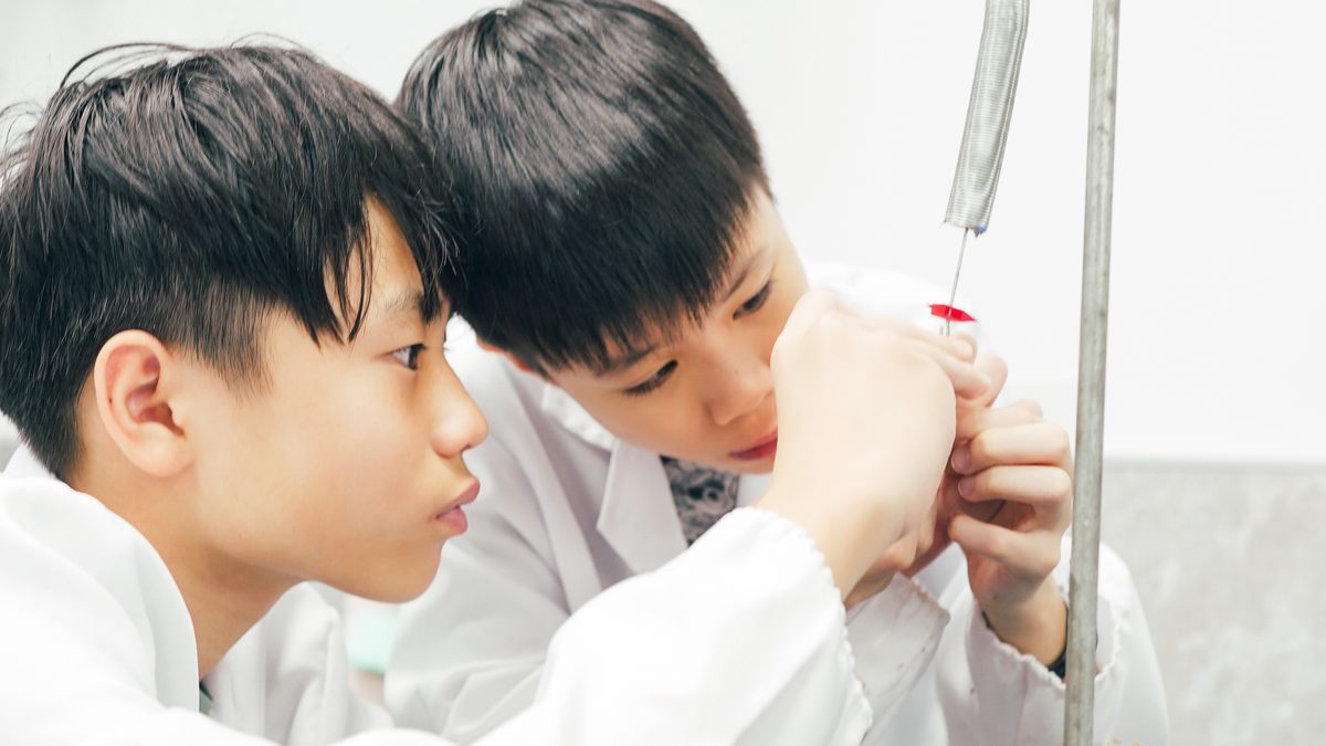 Two junior-high boys in lab coats, heads almost touching, watching the stopcock of a titration set-up on its stand while one of them reaches up to make a fine adjustment.