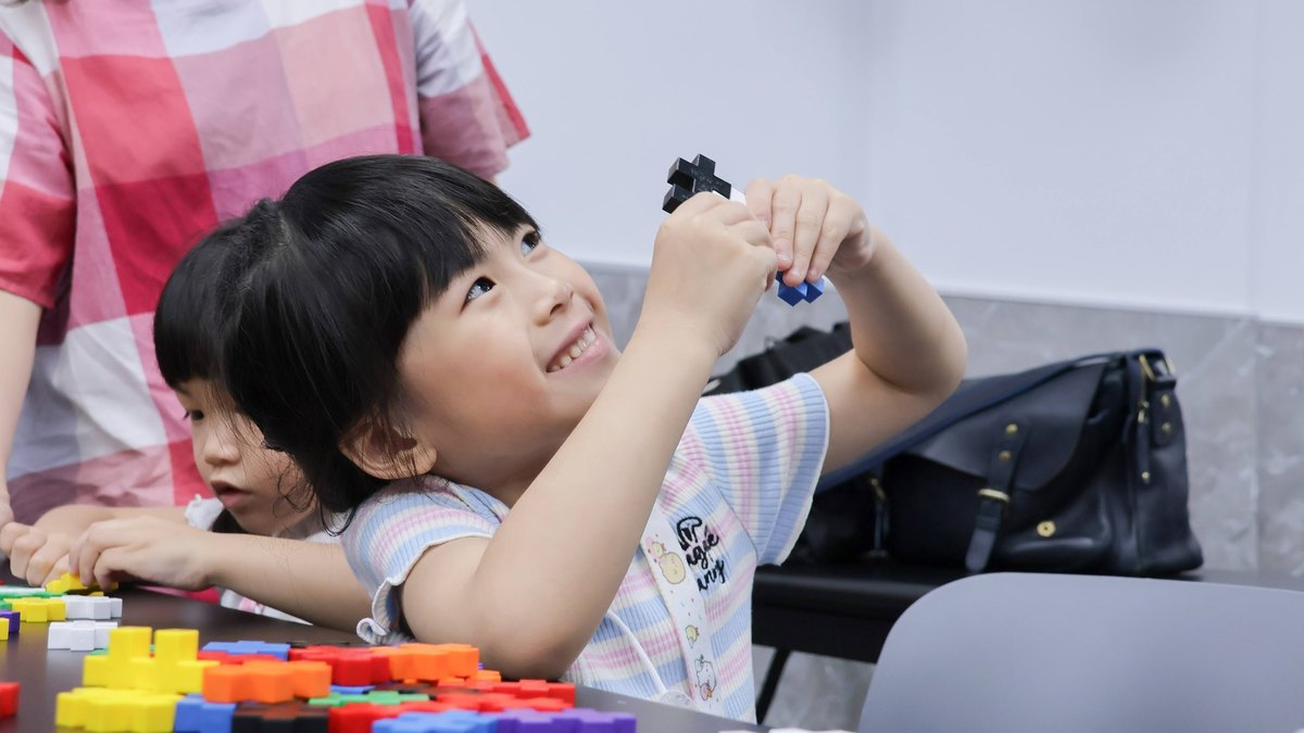 A young child grins while holding two interlocking blocks up to examine them; a scatter of red, yellow and blue building blocks covers the table, with another child reaching in for a piece.