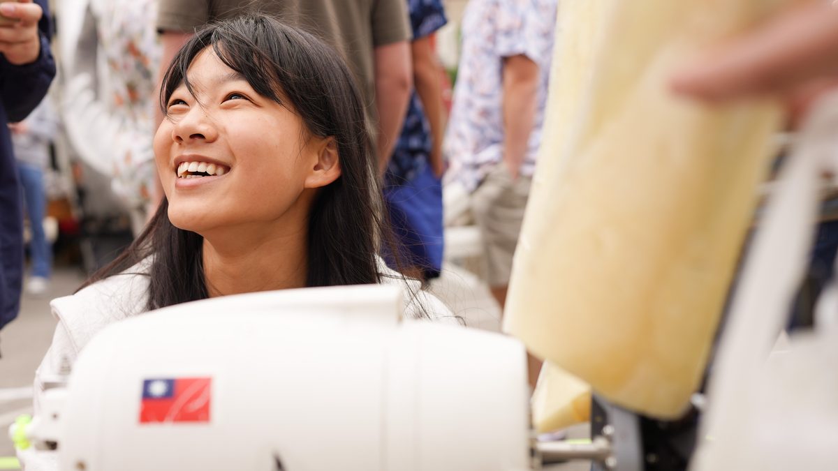 A high-school girl laughing as she looks up, the white nacelle of her self-built wind turbine in front of her with a Taiwanese flag decal on it, and a pale yellow blade at the right of frame.