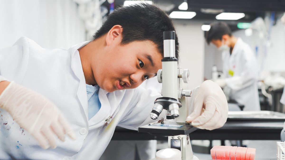 A boy in a lab coat and gloves leaning right into a microscope, one gloved hand adjusting the slide on the stage, his face screwed up in concentration; another student works at their own station behind him.