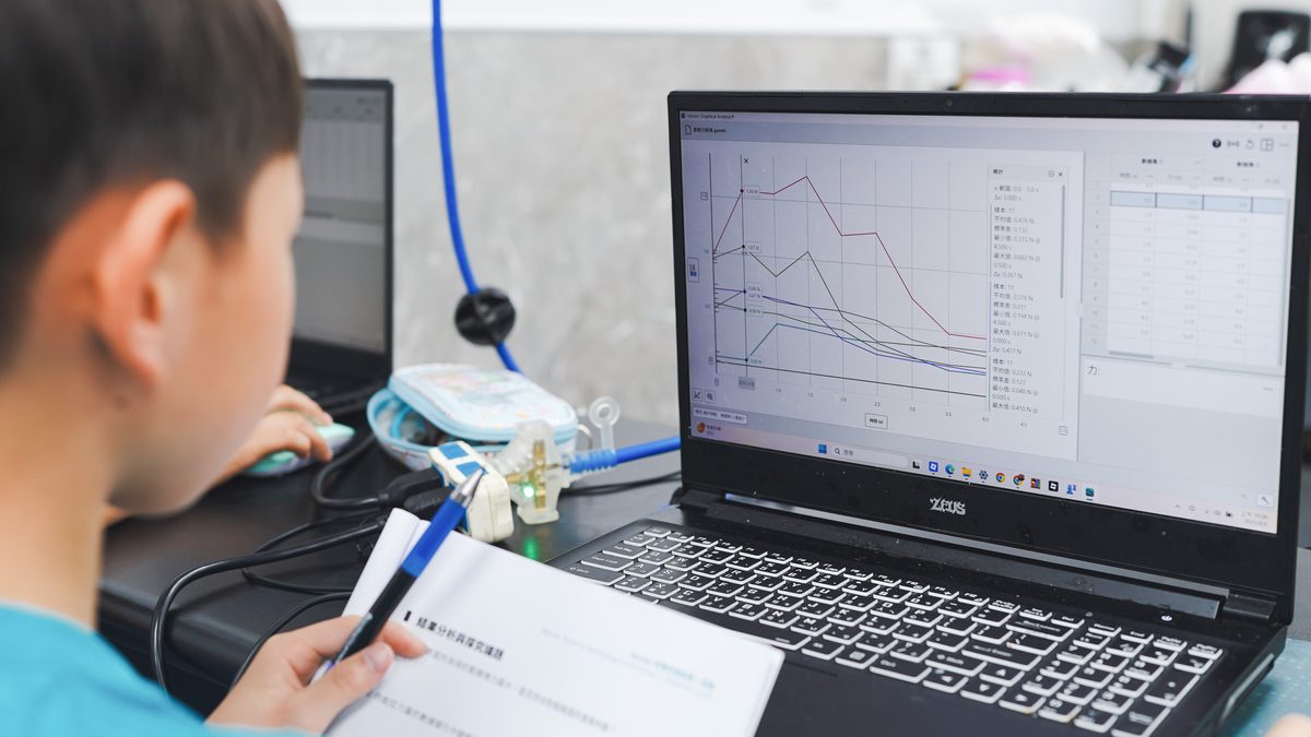 A student holding a pen over an open workbook while looking up at a laptop screen full of live force-measurement curves and a statistics panel, sensors and leads connected on the bench.