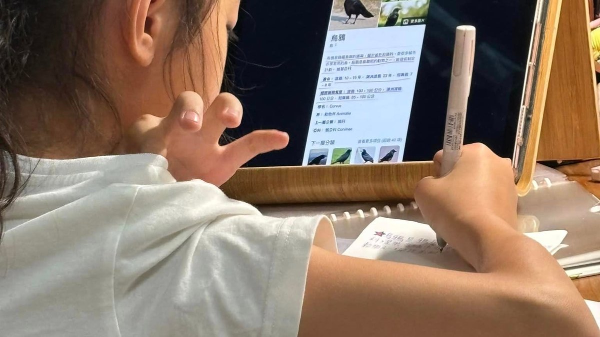 Shot from behind, a child props up a tablet showing an encyclopedia entry on crows — habitat, wingspan, taxonomy — while copying notes into a notebook by hand.