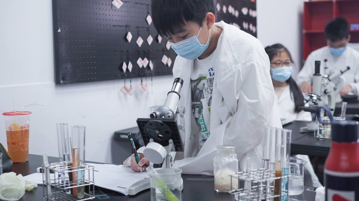 In the lab, a masked student in a white coat leans over a notebook to record results, test-tube racks and a microscope in front of him, two classmates at their own microscopes behind.