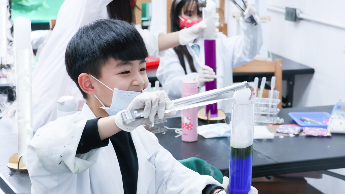 A boy in a white coat and gloves works a pair of tongs into a test tube of layered blue-green liquid, while behind him another student holds up a cylinder of purple solution to inspect it.