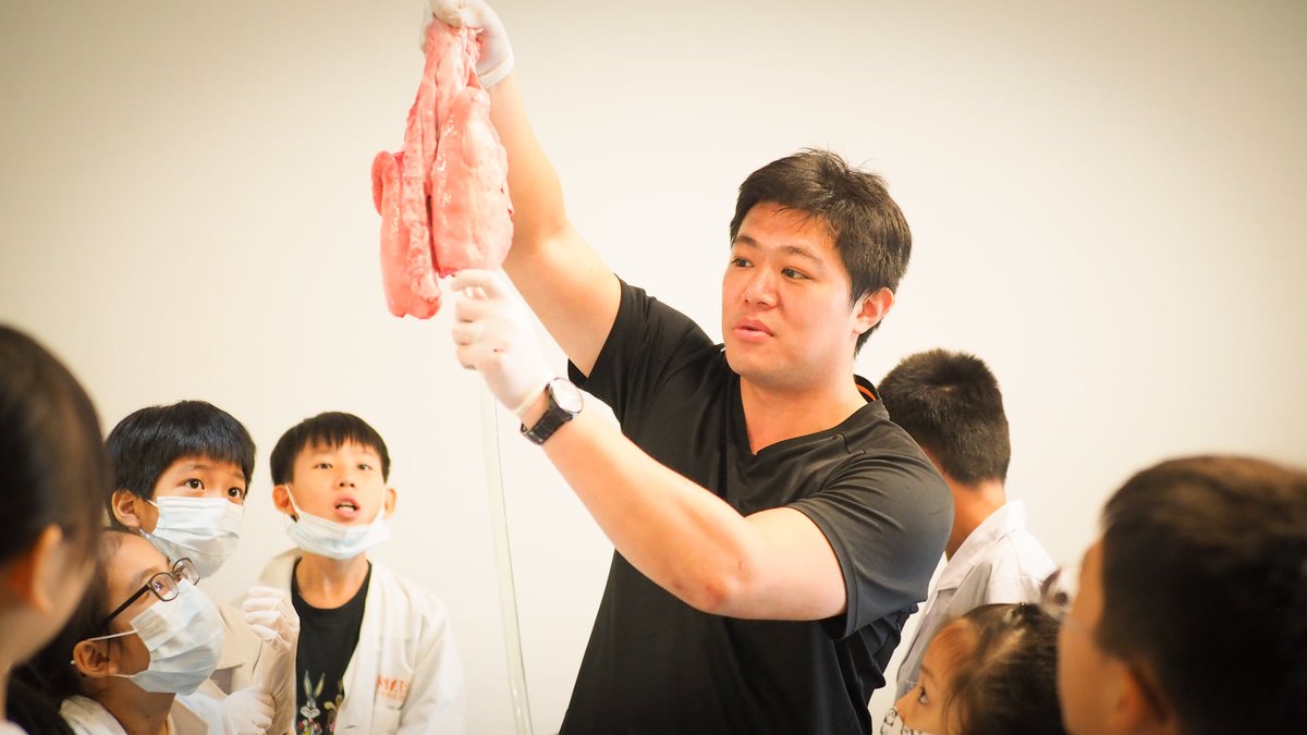 A gloved instructor holds a pair of pig lungs up above the bench while a ring of masked children look up at the specimen.