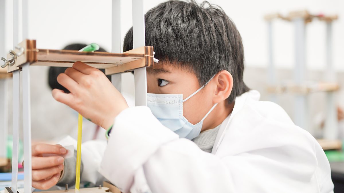 A masked boy in a lab coat squinting closely at the joint of a wooden structure he has built, both hands steadying the beam, with another identical frame behind him.