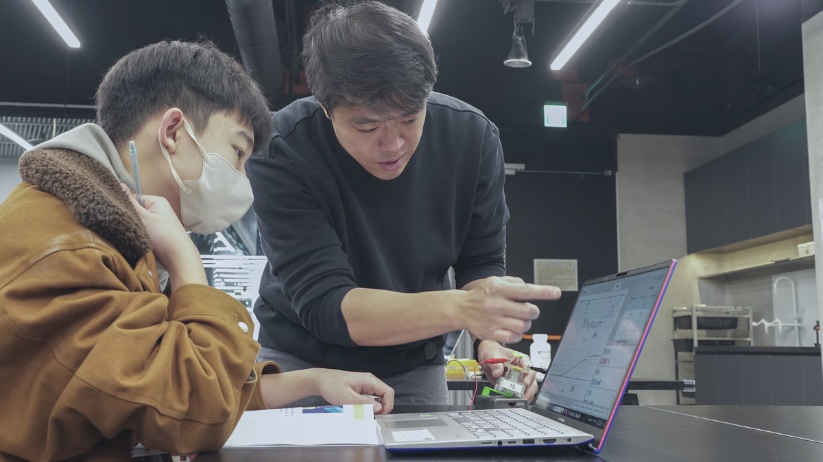 An instructor leans in to point at a live data curve on a laptop screen while a masked student watches, pen in hand; a sensor and its leads sit wired up on the bench.