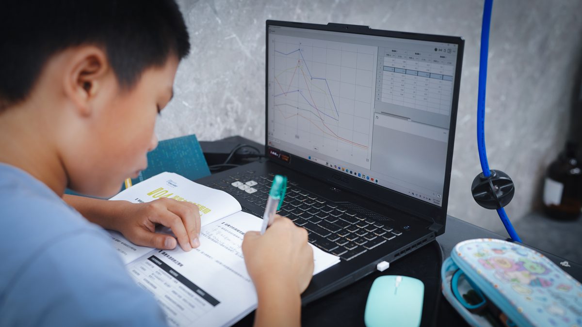 A boy writing in an open workbook, a laptop beside him showing several coloured data curves and a table of readings, with a blue sensor lead running across the bench.